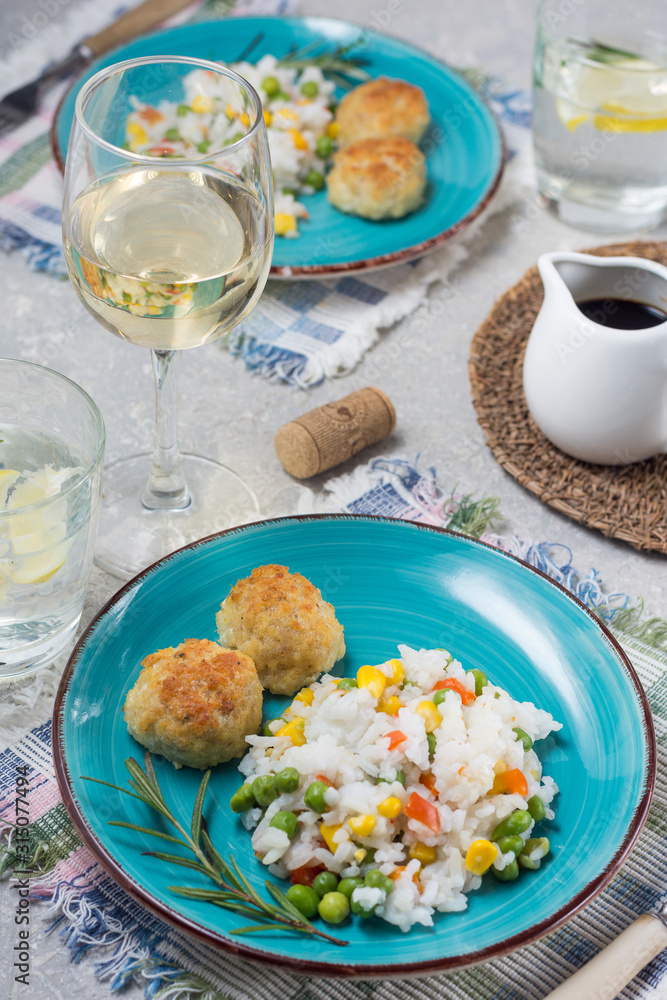 Rice with green peas and corn with fishcakes on bright plates with glasses of water and lemon, as well as a glass of white dry wine, with soy sauce in a sauceboat on a gray table background