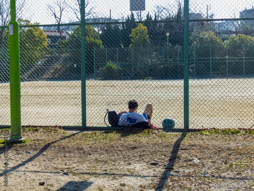 バスケットボールの練習後、グラウンドのフェンスにもたれて休む少年