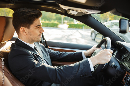 Image of young caucasian businesslike man in black suit driving car