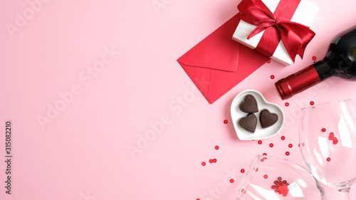 Valentine's day background with wine bottle, gift box, glasses, heart shaped candies, red paper envelope and confetti on pink background. Greeting card template for Valentines Day. Flat lay, top view