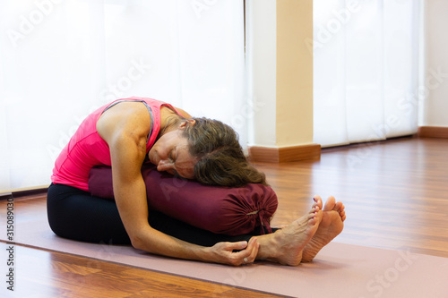 Middle aged woman on pink tank top in paschimottanasana pose resting chest on bolster at bright studio. Female yogi on seated forward bend asana. Therapeutic exercise concept