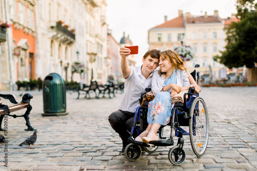 Selfie time. Handsome happy young man hugging his disabled young pretty woman and smiling while taking selfie with her in the city