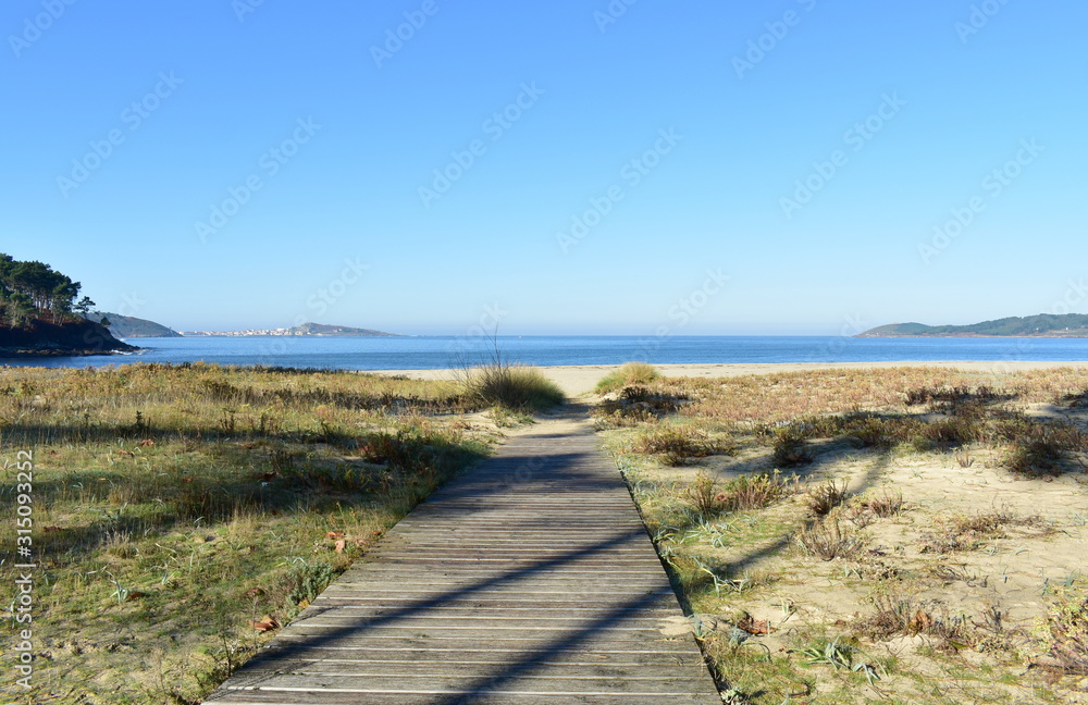 Beach with wooden boardwalk and grass. Bay with blue sea on a sunny day. Muxia, Spain.