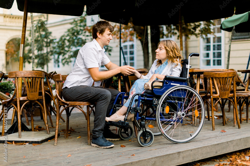 Obraz premium Pretty young woman on a wheelchair enjoying some good company on a date with her handsome man at a cafe outdoors in the city