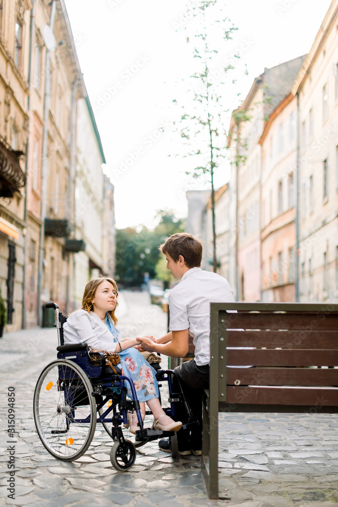 handsome man and pretty woman in wheelchair looking at each other on street. Lovely couple in wheechair walking together in the city