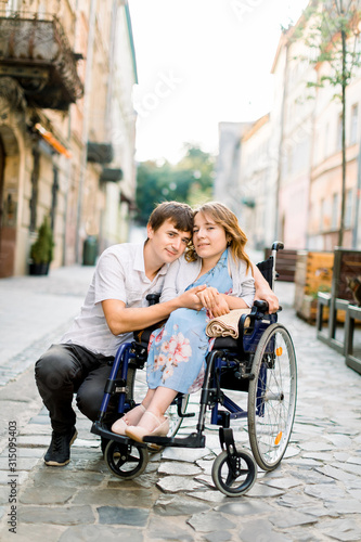 Pretty young smiling woman in the wheelchair and handsome man looking at camera, holding hands, in love, walking in the old city center. © sofiko14