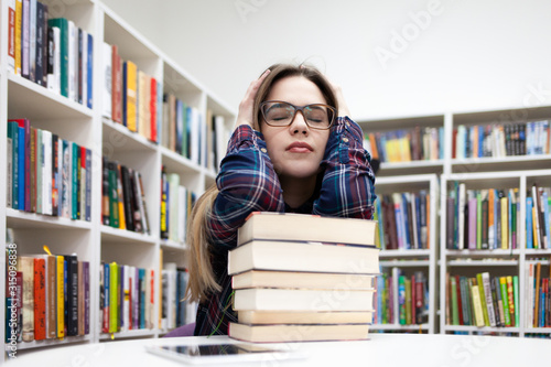 Young tired student girl sitting in a library at a table leaning on a large pile of books. Depressed woman in a plaid shirt prepares for exams in the library