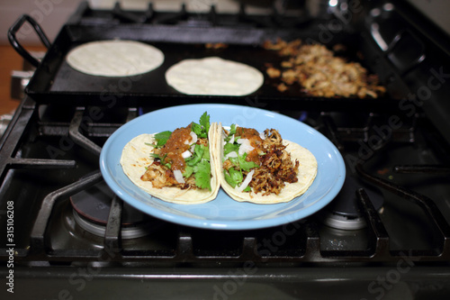 Pork tacos on a plate also known as "carnitas" tacos, on top of a stove with tacos cooking in the background.