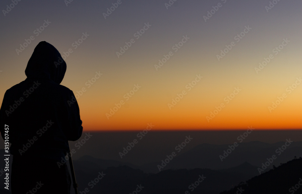 silhouette of Travel photographer standing with a camera mounted on a tripod and shooting a time lapse of the sunrise/sunset. man wearing his hood enjoying the mountain view /valley view