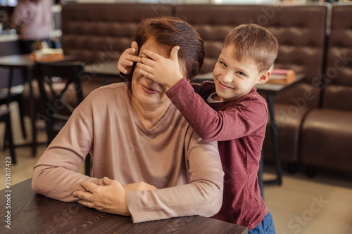grandmother and grandson spend time together in a cafe. grandson prepares a surprise for her grandmother and closes her eyes with her children's hands