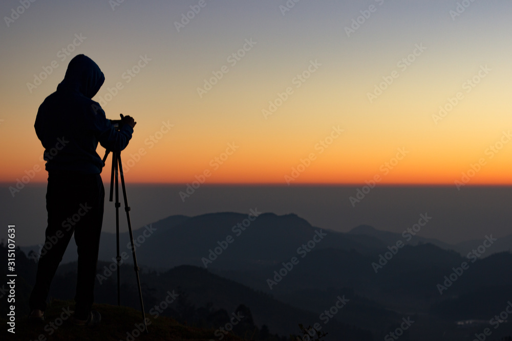 silhouette of Travel photographer standing with a camera mounted on a tripod and shooting a time lapse of the sunrise/sunset. man wearing his hood enjoying the mountain view /valley view