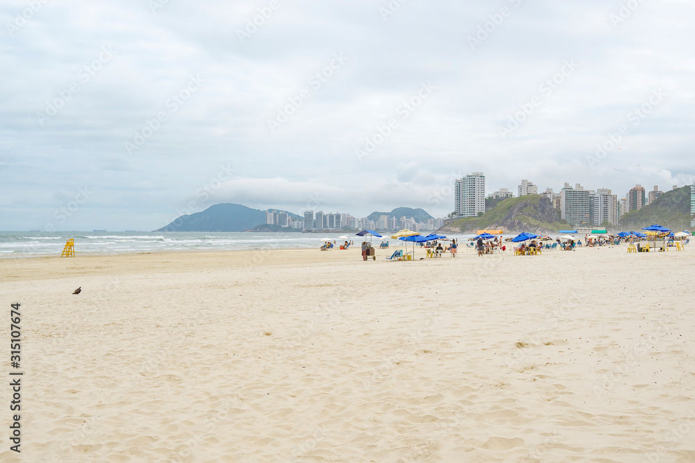 People enjoying the day at Enseada beach - Guaruja SP Brazil. Cloudy day, sunshades on the sand, tourists, in front of the sea. Praia da Enseada.