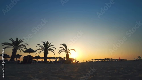 Woman in a hat walking on the sand and watching sunset at tropical beach with palm trees