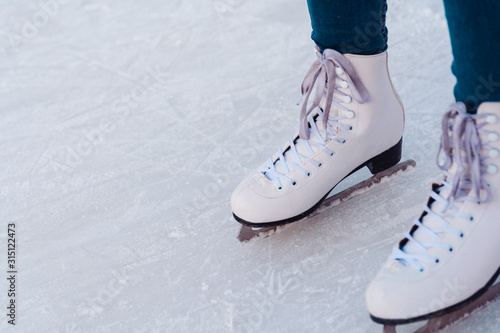 A young woman in white figure skates and blue jeans is standing on the ice, ready for the ride on the rink. Training. Winter entertainment and pastime. Leisure and lifestyle. View from above. Closeup.