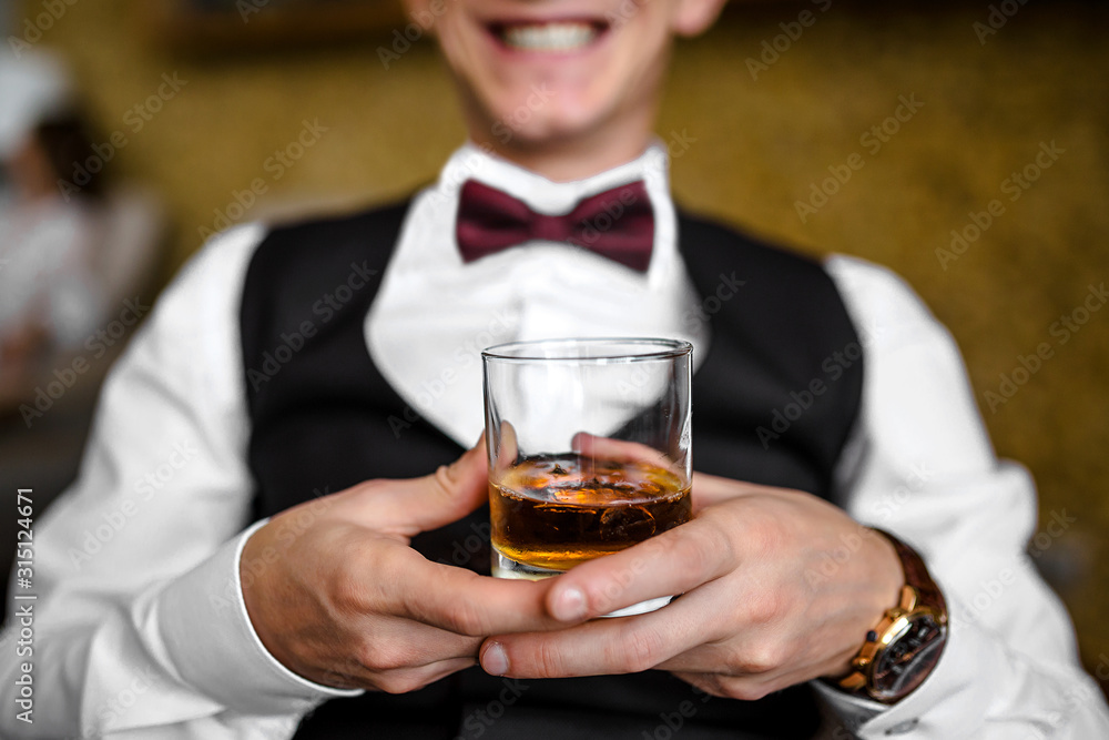 Handsome groom in white shirt sitting in luxury leather chair and holding whiskey morning before the wedding, confident business man holding cognac in glass while resting in armchair