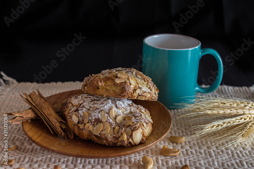 Traditional French croissant with almonds on the wood plate and a green mug with a beverage on dark background