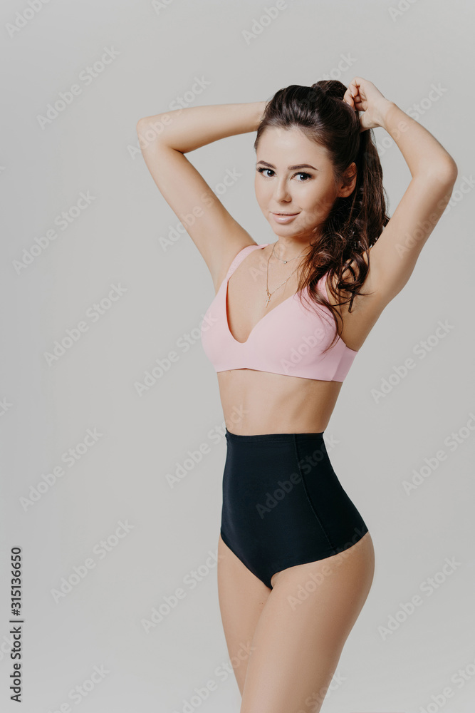Obraz premium Vertical shot of sexy lady in sexy bra and black pants, has slim perfect figure, dark hair combed in pony tail, models against grey background, looks straightly at camera, has beautiful body