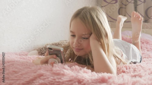 Cute girl playing smartphone lying on her bed in her room.