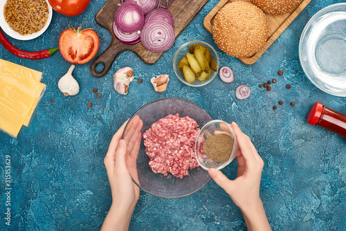top view of woman adding black pepper to minced meat near burger ingredients on blue textured surface