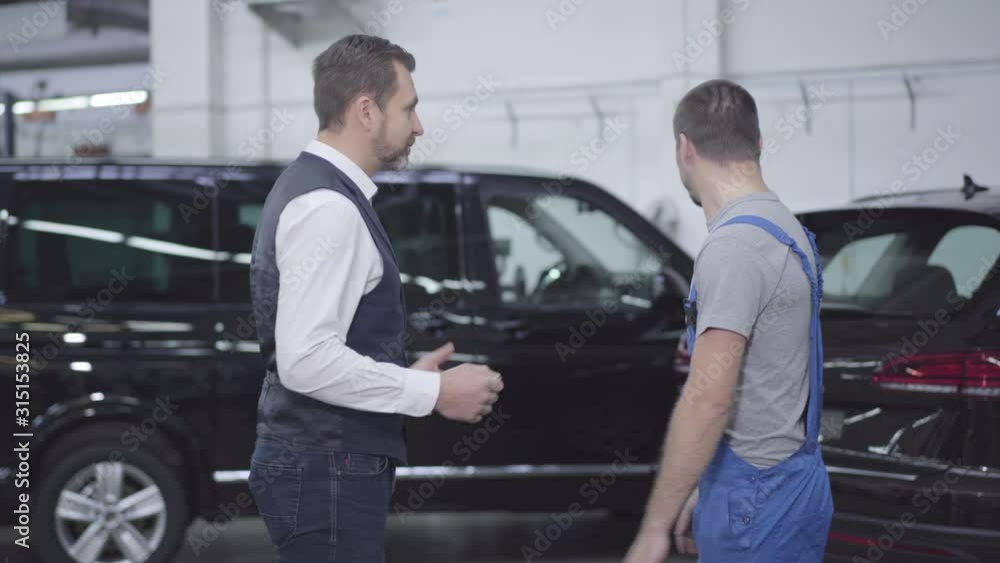 Side view portrait of adult Caucasian businessman standing in car repair shop and talking with worker. Handsome successful man consulting with auto mechanic regarding his automobile. Repair services. Side view portrait of adult Caucasian businessman standing in car repair shop and talking with worker. Handsome successful man consulting with auto mechanic regarding his automobile. Repair services.