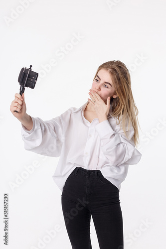 happy girl makes self portrait with smartphone attached to small tripod. young pretty blond female with long hair sending air kiss, using cell phone isolated on white background. teen communication