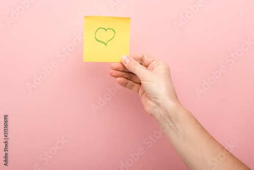 cropped view of woman holding sticky note with heart on pink background
