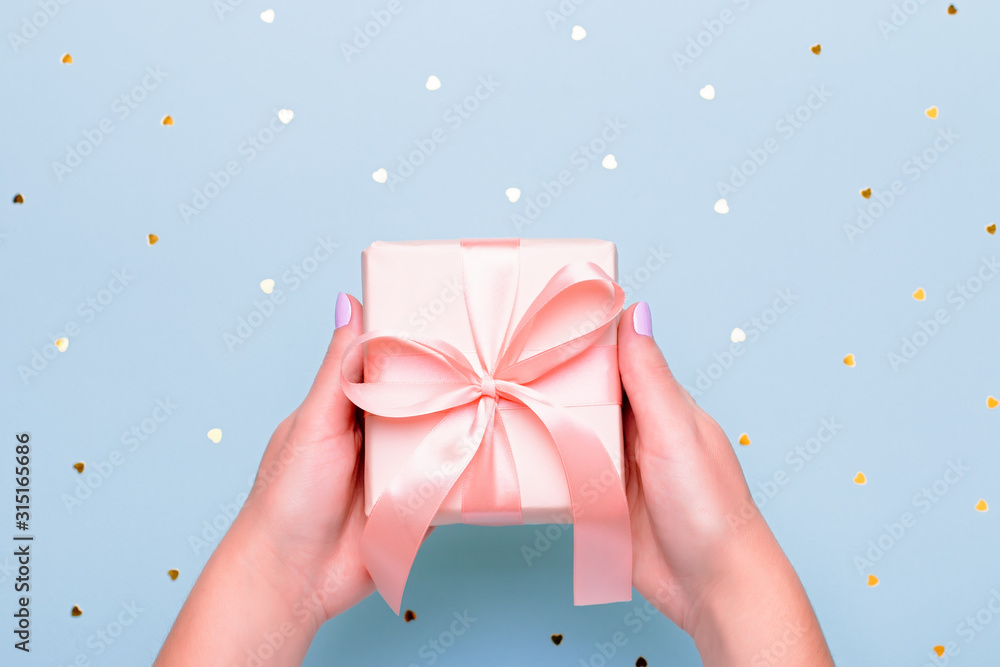 Woman hands holding gift box in pink color on pastel blue background with heart shape confetti, copyspace. Valentine's Day greeting card in trendy colors, Happy Valentine Day background Woman hands holding gift box in pink color on pastel blue background with heart shape confetti, copyspace. Valentine's Day greeting card in trendy colors, Happy Valentine Day background