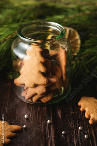 Gingerbread cookies Christmas fir-tree and heart shape in shiny glass jar on wooden background, citrus dried lemon, fir tree branch, angle view, selective focus