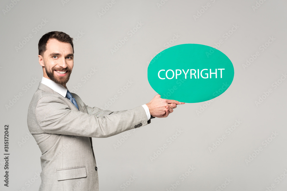 handsome businessman holding speech bubble with word copyright and smiling at camera isolated on grey handsome businessman holding speech bubble with word copyright and smiling at camera isolated on grey