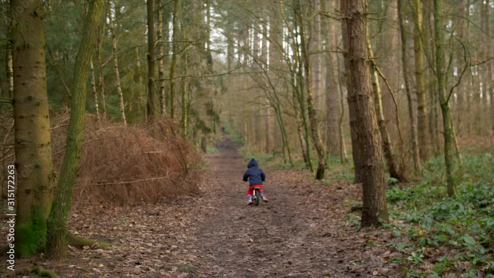 Cute Toddler Riding His Bike Through The Woods In British Park, Muddy Path Exploring And Adventuring. Tall Trees Surrounding. Having Fun On His Bike. Camera Tracking Him. Cute Toddler Riding His Bike Through The Woods In British Park, Muddy Path Exploring And Adventuring. Tall Trees Surrounding. Having Fun On His Bike. Camera Tracking Him.