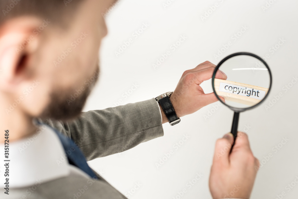 selective focus of businessman looking at word copyright on wooden block through magnifying glass isolated on grey selective focus of businessman looking at word copyright on wooden block through magnifying glass isolated on grey