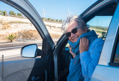 A smiling senior woman wears the scarf near her face. Casual dress. Wind turbines in the background. Clean energy concept