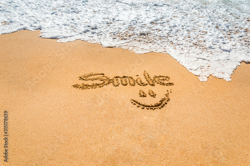 Hand written text smile and symbol on the golden beach sand with coming wave