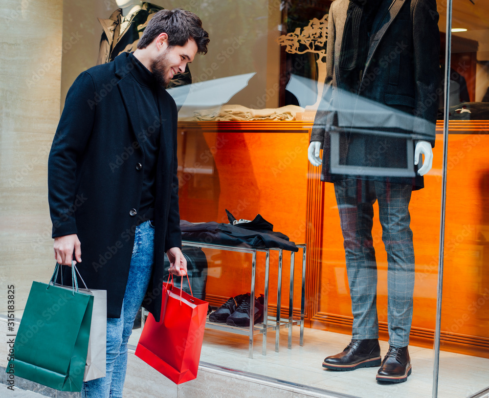 Young attractive bearded man with bags goes shopping in the city and looks at the shop window laughing.