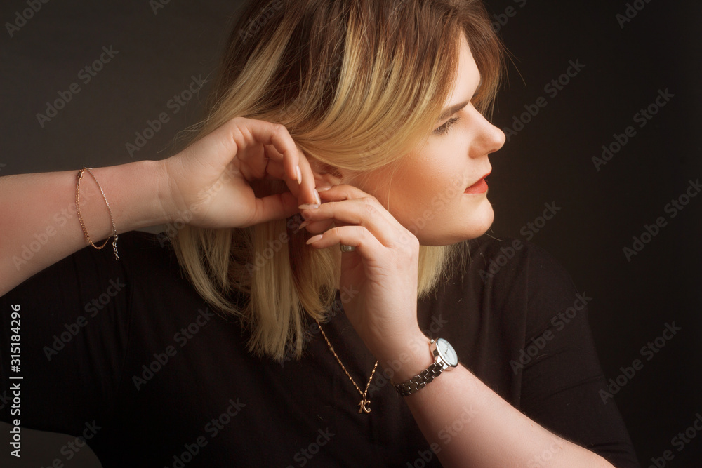blonde Plus Size Model on a black background in dark clothes, fastens the earring with two hands