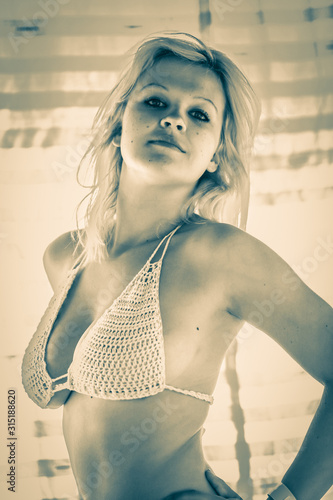 Young attractive blond woman in bikini posing on the bed. Sexy lady with big boobs and knitted bikini bathing suit posing in bedroom with long white curtains and blinds looking at the camera smiling photo