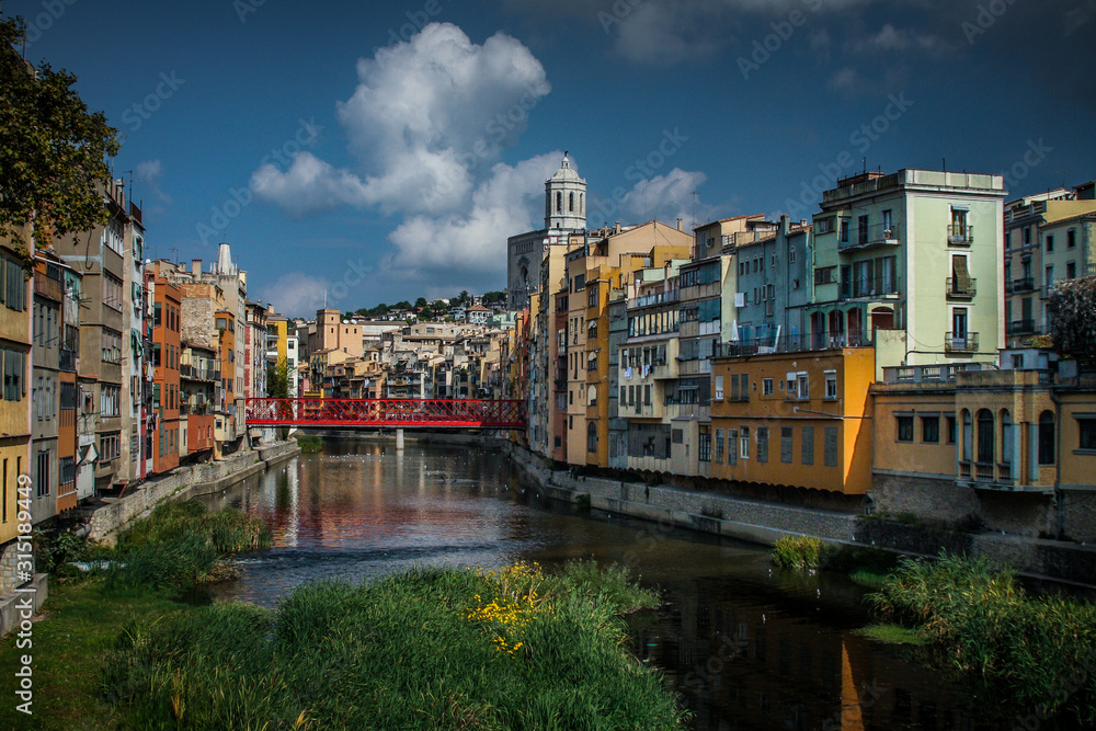 Panorama of Girona from the river, with Girona cathedral of Saint Mary and red Eiffel bridge seen in the foreground. Greenery on the water is seen.
