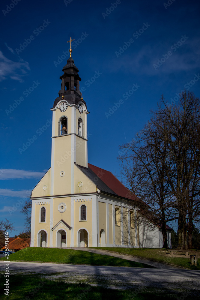 Typical central european church with belltower in white and yellow color on a top of the hill of Urh in Ljubljana, Slovenia during the late spring afternoon.