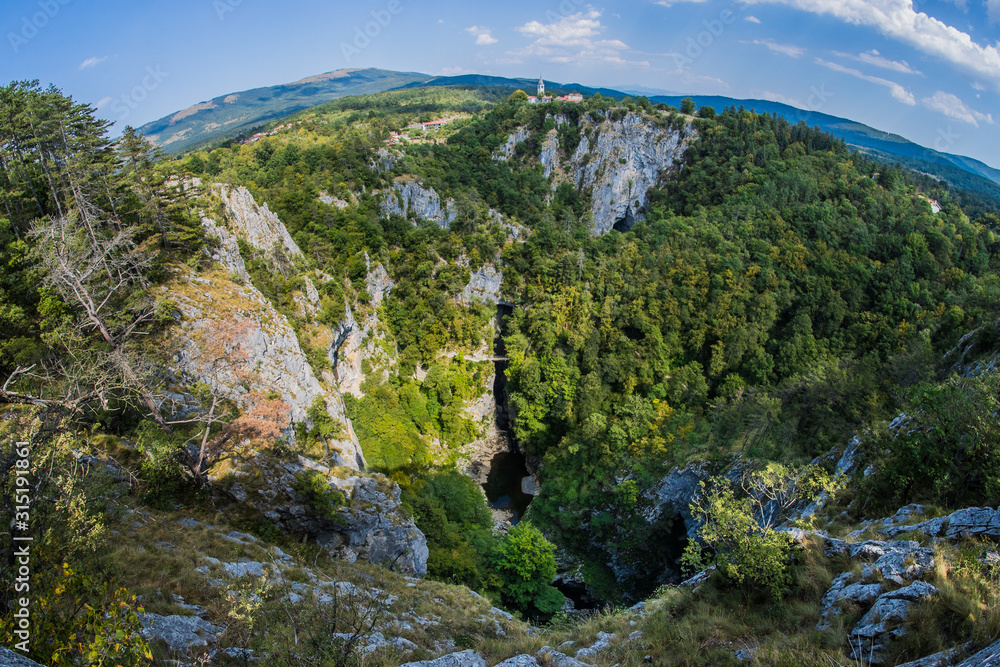 Unesco world heritage site Skocjanske jame taken with a wide fish eye view. Looking towards the deep gorge with cave entrance and walking paths. Village of Skocjan in the background.