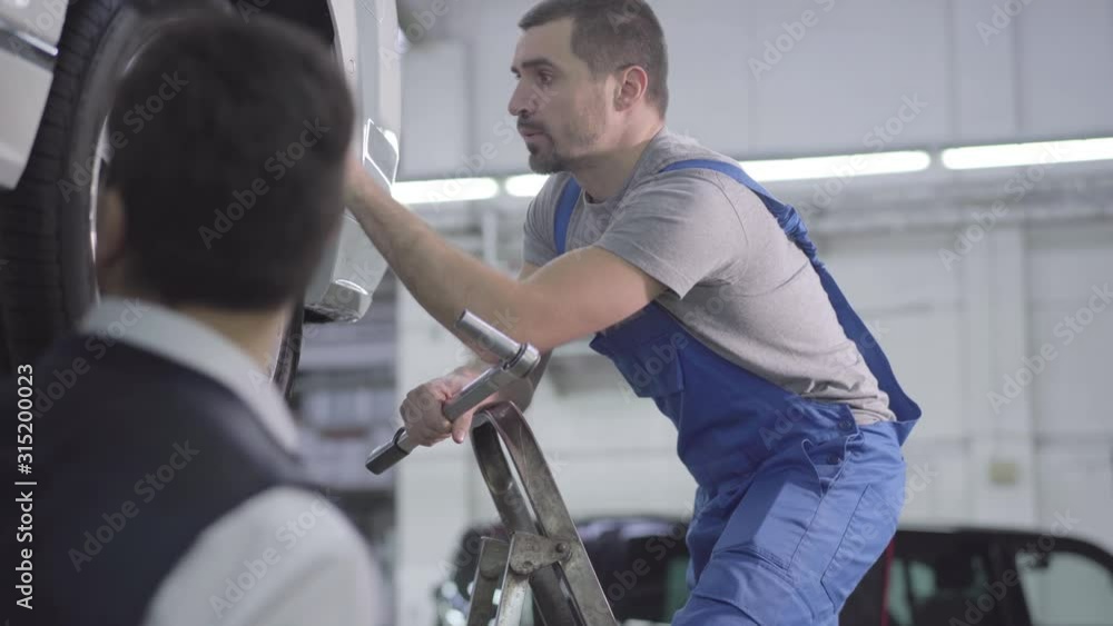 Close-up of two Caucasian men next to broken car talking. Young repair man in blue robe holding adjustable wrench and explaining problem to customer. Car repair, repair shop. Close-up of two Caucasian men next to broken car talking. Young repair man in blue robe holding adjustable wrench and explaining problem to customer. Car repair, repair shop.