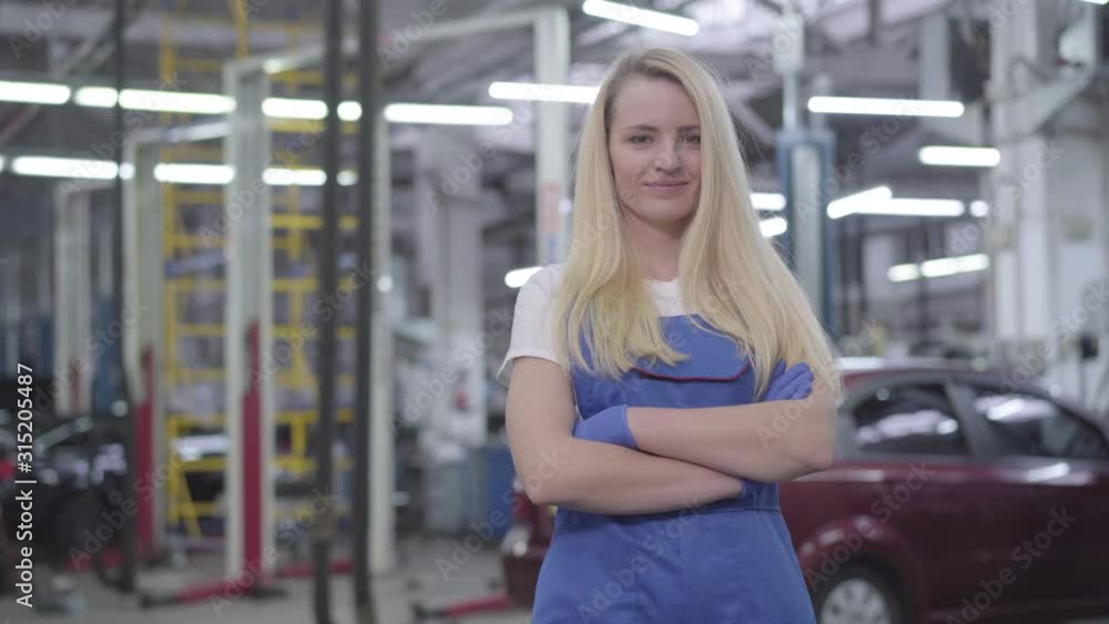 Portrait of blond confident Caucasian woman in blue workrobe standing in repair shop, looking at camera and smiling. Attractive female auto mechanic at workplace. Occupation, professional. Portrait of blond confident Caucasian woman in blue workrobe standing in repair shop, looking at camera and smiling. Attractive female auto mechanic at workplace. Occupation, professional.