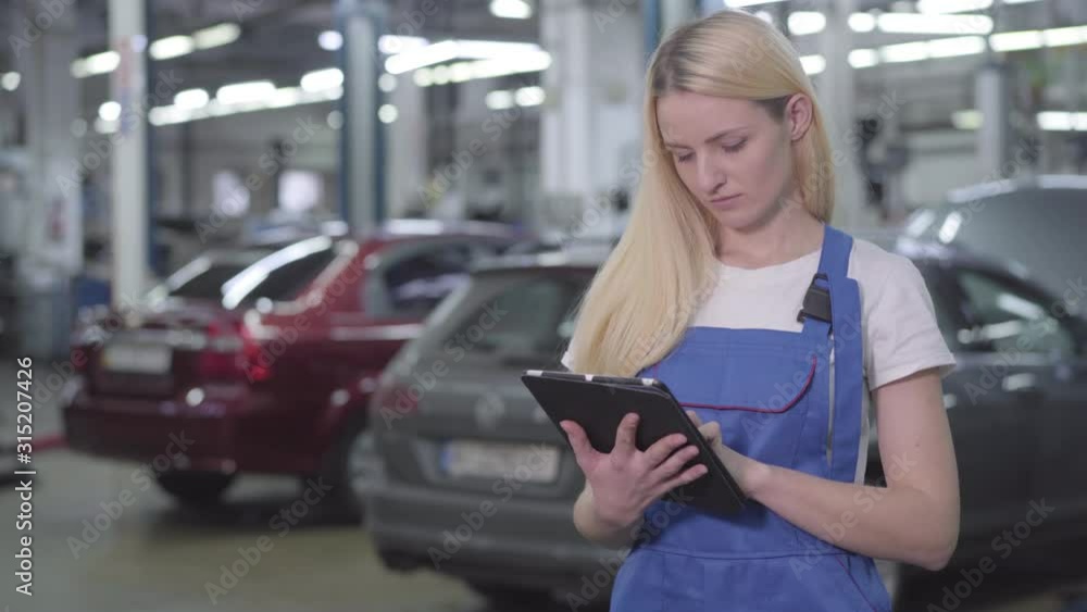 Portrait of young Caucasian woman in workrobe holding tablet and smiling. Beautiful professional auto mechanic standing in car repair shop. Professional, warranty services. Portrait of young Caucasian woman in workrobe holding tablet and smiling. Beautiful professional auto mechanic standing in car repair shop. Professional, warranty services.