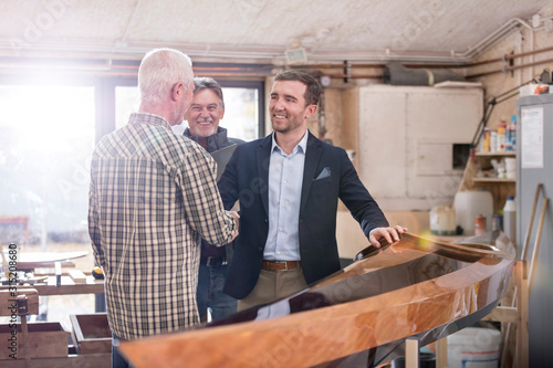 Male carpenters handshaking satisfied customer next to wood kayak in workshop