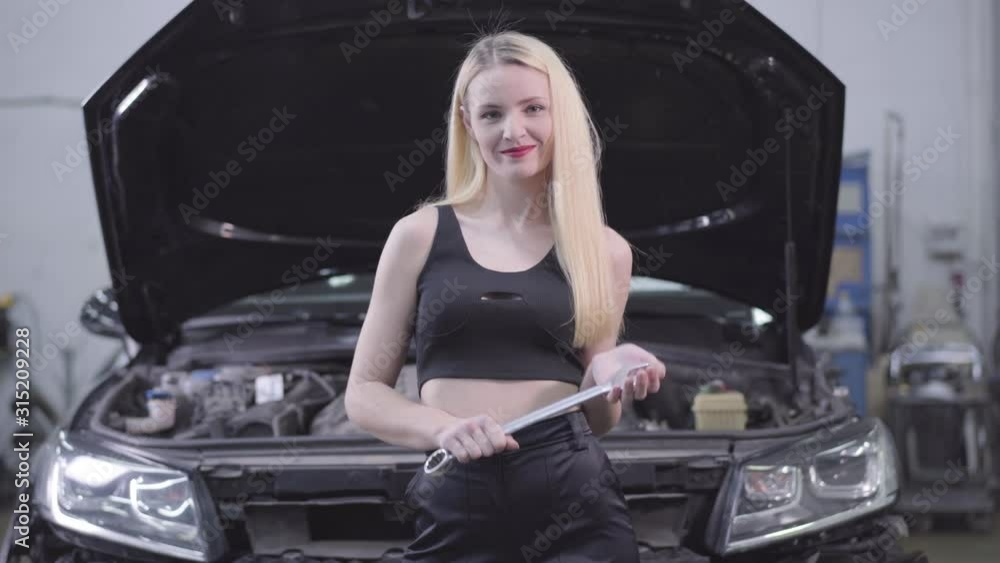 Sexy blond Caucasian woman standing at the background of open car hood with adjustable wrench and smiling. Beautiful female auto mechanic standing in repair shop. Sexy blond Caucasian woman standing at the background of open car hood with adjustable wrench and smiling. Beautiful female auto mechanic standing in repair shop.