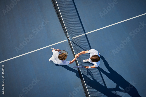 Overhead view young male tennis players handshaking at net on sunny blue tennis court