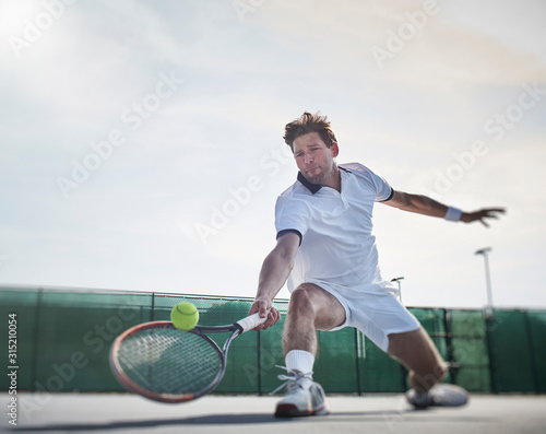 Determined young male tennis player playing tennis, reaching for the ball on sunny tennis court