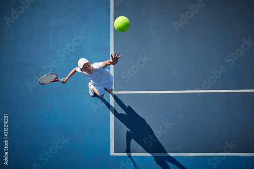 Overhead view of young male tennis player playing tennis, serving the ball on sunny blue tennis court