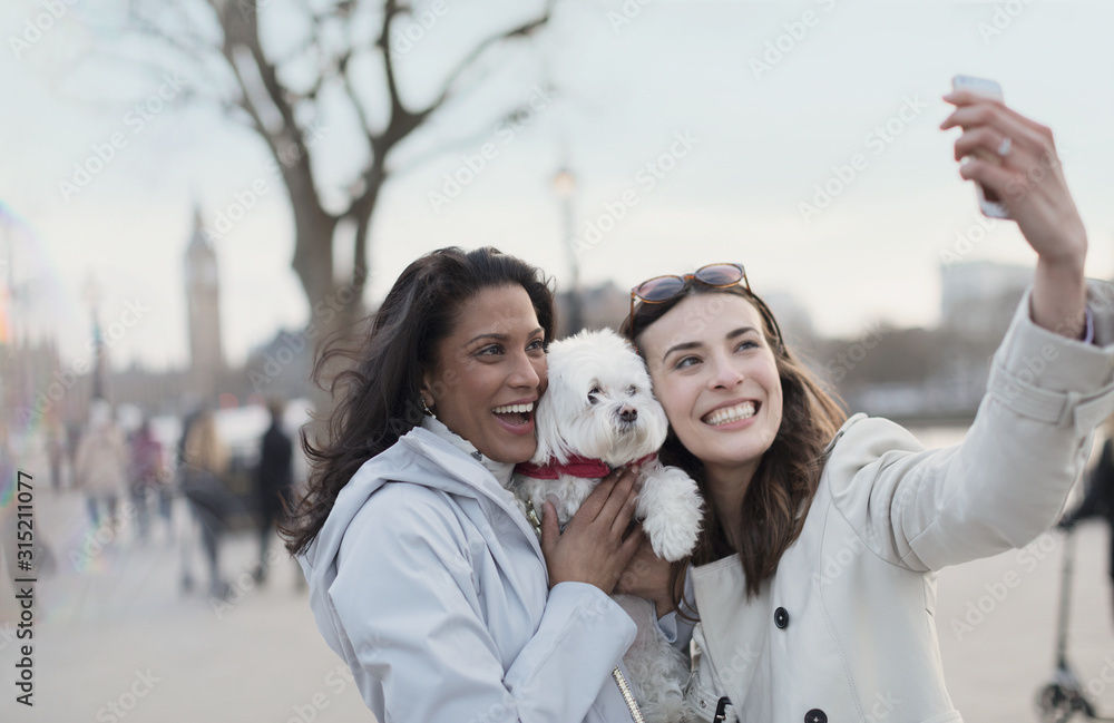 Playful, smiling lesbian couple white dog taking selfie camera phone in urban park, London, UK Playful, smiling lesbian couple white dog taking selfie camera phone in urban park, London, UK