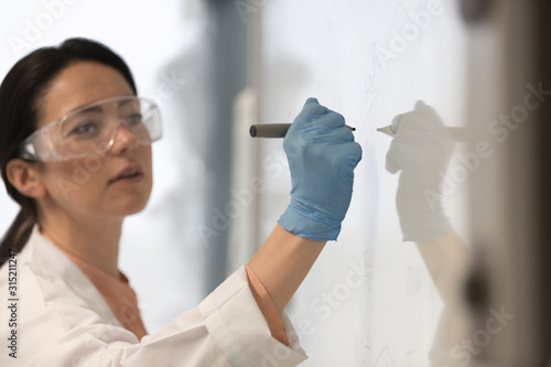 Female science teacher wearing lab coat, rubber glove and goggles, writing at whiteboard in classroom