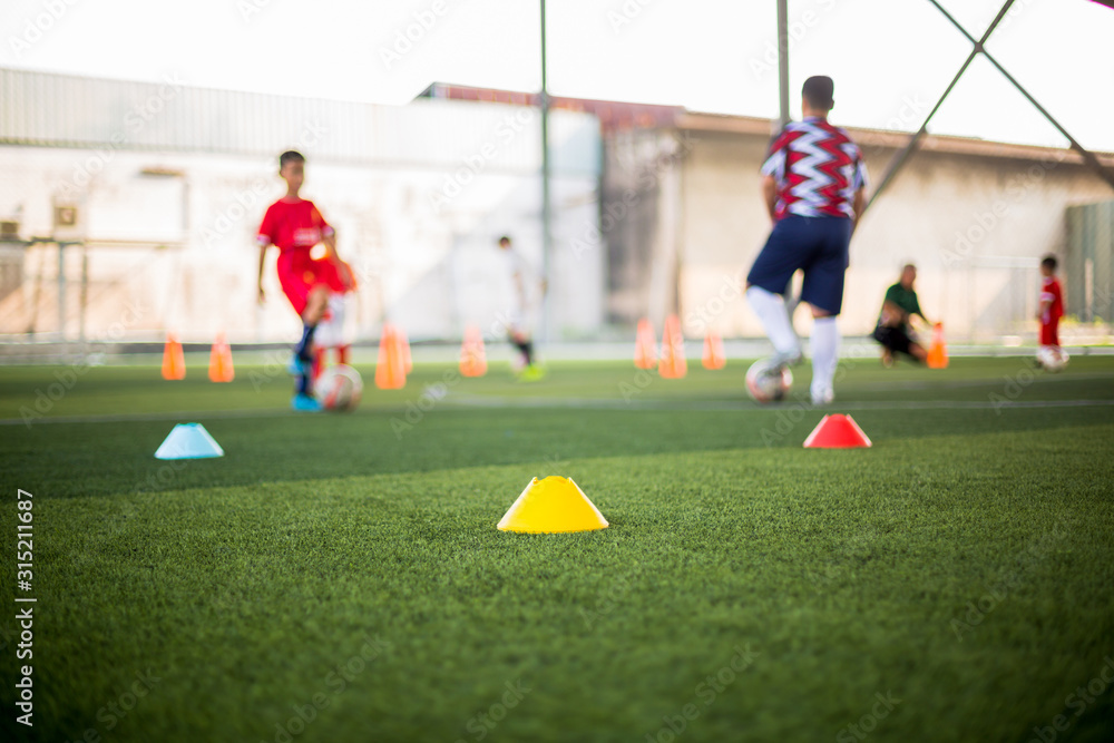 Obraz premium Marker Cone on green artificial turf with blurry kid soccer players are training background. Soccer academy.